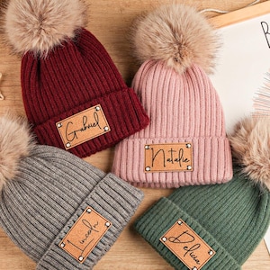 May include: Four personalized knit beanies in burgundy, pink, gray, and green, each with a faux fur pom-pom. Each beanie has a leatherette patch with a name. A decorative sign with a lion illustration and the words "BE BRAVE" is in the background.