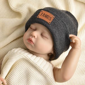 May include: A sleeping baby wearing a gray beanie with a brown leather patch that says "JAMES" on it.