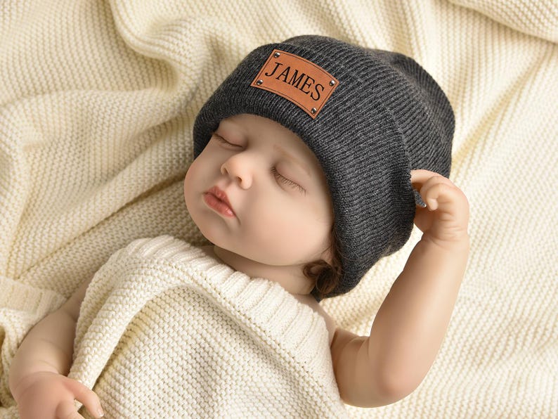 May include: A sleeping baby wearing a gray beanie with a brown leather patch that says "JAMES" on it.