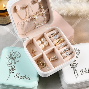 May include: Three small, square jewelry boxes with floral designs and personalized names. The boxes are white, light blue, and pink. The pink box is open and shows compartments for rings, earrings, and a necklace.