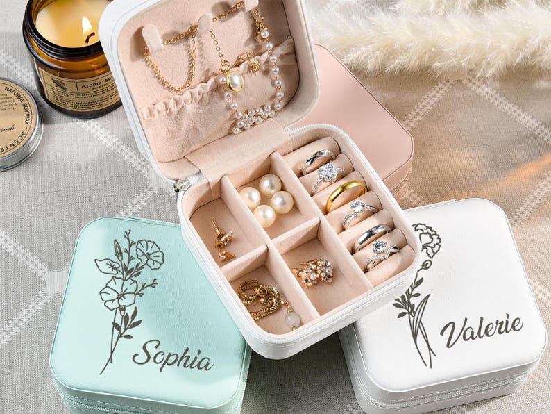 May include: Three small, square jewelry boxes with floral designs and personalized names. The boxes are white, light blue, and pink. The pink box is open and shows compartments for rings, earrings, and a necklace.