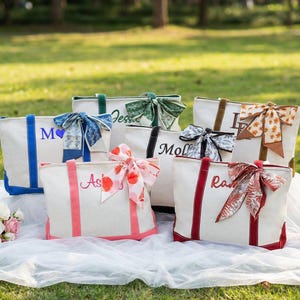 May include: A collection of personalized tote bags in various colors, each adorned with a decorative bow and a name embroidered on the front. The bags are arranged on a white cloth outdoors, with a bouquet of flowers and a bottle of champagne nearby.