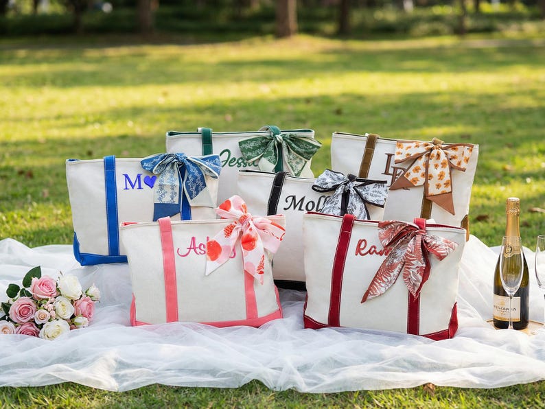 May include: A collection of personalized tote bags in various colors, each adorned with a decorative bow and a name embroidered on the front. The bags are arranged on a white cloth outdoors, with a bouquet of flowers and a bottle of champagne nearby.