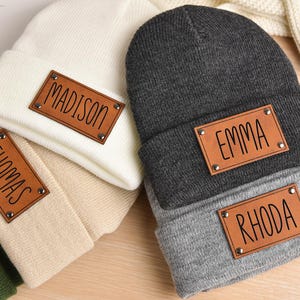 May include: Four knit beanies with personalized leather patches. The beanies are in beige, gray, black, and green. The patches have the names Madison, Thomas, Emma, and Rhoda.