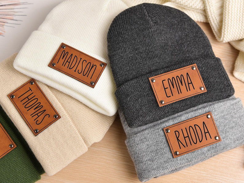 May include: Four knit beanies with personalized leather patches. The beanies are in beige, gray, black, and green. The patches have the names Madison, Thomas, Emma, and Rhoda.