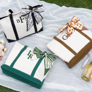 May include: Three canvas tote bags with personalized initials and decorative bows. One bag has black trim and a black and white bow, another has brown trim and a floral bow, and the third has green trim and a green bow. A bouquet of pink and white roses and a bottle of champagne are also present.