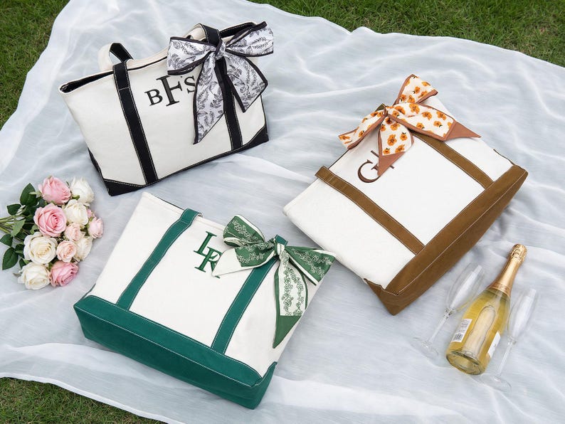 May include: Three canvas tote bags with personalized initials and decorative bows. One bag has black trim and a black and white bow, another has brown trim and a floral bow, and the third has green trim and a green bow. A bouquet of pink and white roses and a bottle of champagne are also present.