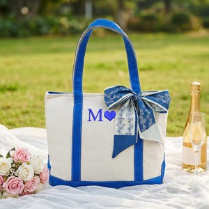 May include: A white tote bag with blue accents, handles, and a bow. The bag features the letter "M" and a blue heart. A bouquet of pink and white roses, champagne, and two glasses are also present.