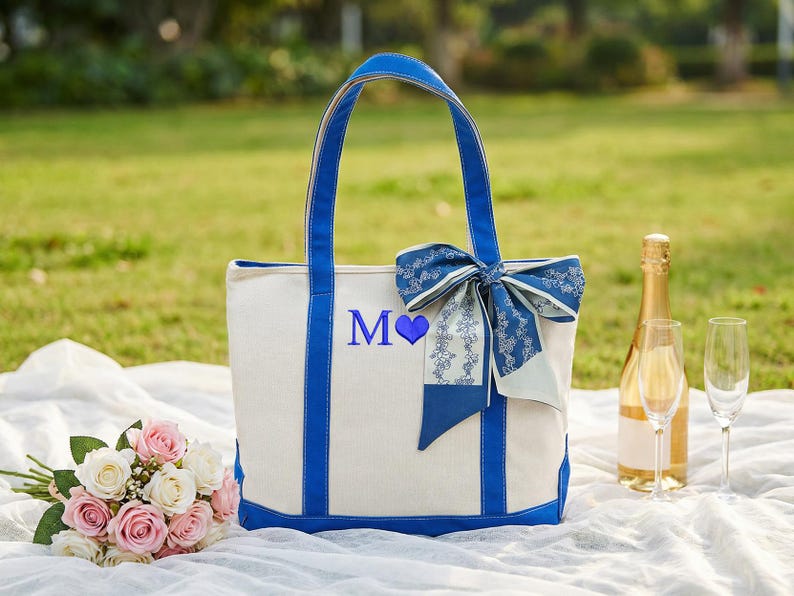 May include: A white tote bag with blue accents, handles, and a bow. The bag features the letter "M" and a blue heart. A bouquet of pink and white roses, champagne, and two glasses are also present.