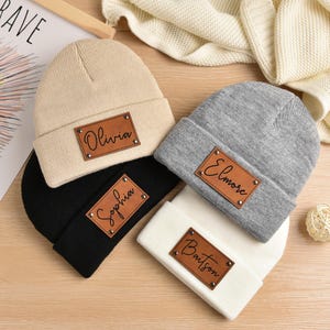 May include: Four knitted beanies in different colors with personalized leather patches. The patches have the names Olivia, Sophia, Elmore, and Baitson written in cursive.