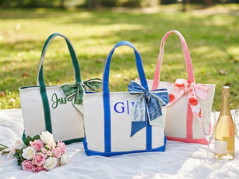 May include: Three canvas tote bags with colorful accents. One bag has green handles and trim, with the name "Jessica" embroidered in green. Another has blue handles and trim, with the initials "G|I" embroidered in blue. The third bag has pink handles and trim.