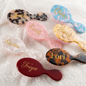 Könnte beinhalten: Eine Sammlung von sieben ovalen Haarbürsten in verschiedenen Farben, darunter Schildpatt, Blau, Rosa, Gold, Weiß und Kastanienbraun. Jede Bürste ist mit einem personalisierten Namen in goldener Schreibschrift versehen, wie Amelia, Claire, Karla, Bailey, Sophia, Lori und Freya.