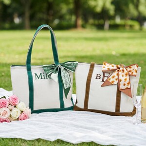 May include: Two canvas tote bags with personalized initials, one green and one brown, sit on a white blanket. The green bag has the name "Mike" embroidered on it. A bouquet of pink and white roses and a bottle of champagne with two glasses complete the picnic scene.