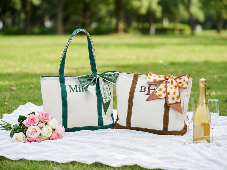 May include: Two canvas tote bags with personalized initials, one green and one brown, sit on a white blanket. The green bag has the name "Mike" embroidered on it. A bouquet of pink and white roses and a bottle of champagne with two glasses complete the picnic scene.