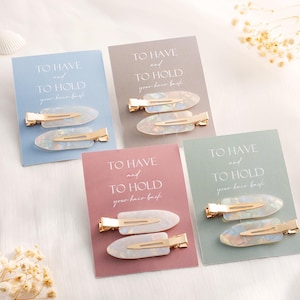 Puede incluir: Cuatro juegos de dos horquillas para el pelo con cierres de metal dorado y detalles acrílicos ovalados, iridiscentes y veteados. Cada par está montado sobre una tarjeta rectangular azul, gris, rosa o verde con el texto TO HAVE and TO HOLD your hair back.