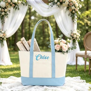 May include: A cream-coloured canvas tote bag with light blue trim and handles. The name Chloe is embroidered in blue script on the front. The bag features a rectangular front pocket and contains rolled newspapers and a bouquet of pink and white flowers.