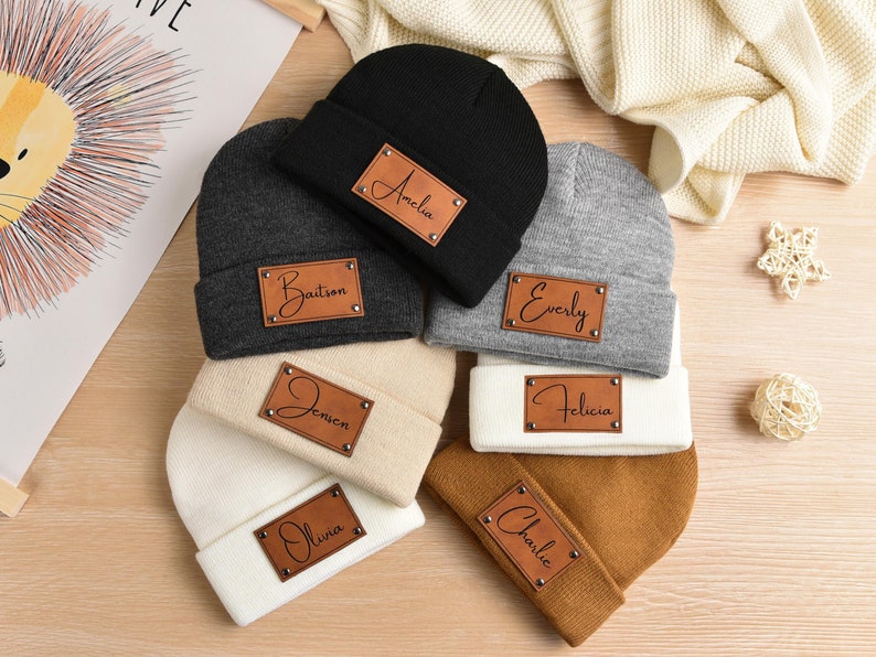 May include: Six different colored knit beanies with personalized leather patches. The patches have the names Amelia, Baitson, Evely, Felicia, Jensen, Olivia, and Charlie written in cursive.