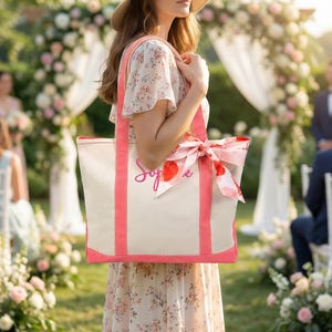 May include: A canvas tote bag with coral trim and handles, featuring the name "Sophie" in cursive script. The bag is adorned with a matching coral and floral patterned scarf tied in a bow. The bag is cream-colored and is being held outdoors.