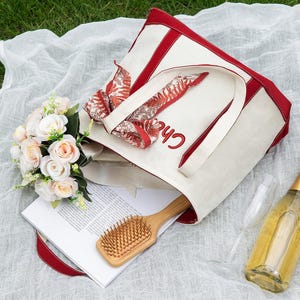 May include: A picnic scene with a cream-colored tote bag with red trim and the name "Chloe" embroidered in red. The bag contains a floral scarf, a wooden hairbrush, and a magazine. A bouquet of roses, a champagne bottle, and two glasses are also present.