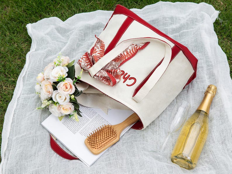 May include: A picnic scene with a cream-colored tote bag with red trim and the name "Chloe" embroidered in red. The bag contains a floral scarf, a wooden hairbrush, and a magazine. A bouquet of roses, a champagne bottle, and two glasses are also present.