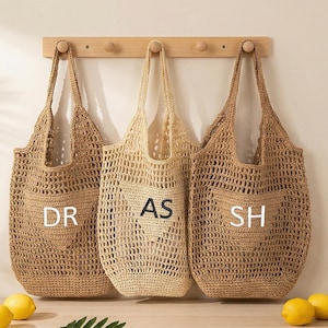 May include: Three open-weave tote bags in shades of beige hang from a wooden rack. Each bag has initials: "DR", "AS", and "SH". The bags are made of a natural fibre and have a pocket on the front. Lemons and bananas are in the foreground.