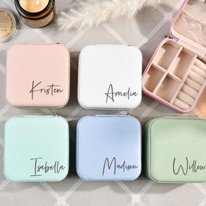 May include: Six small square jewelry boxes with different colored lids. The lids are pink, white, light blue, light green, and two are a darker green. Each lid has a name written on it in black script: Kristen, Amelia, Isabella, Madison, and Willow.