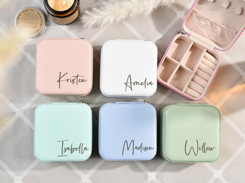May include: Six small square jewelry boxes with different colored lids. The lids are pink, white, light blue, light green, and two are a darker green. Each lid has a name written on it in black script: Kristen, Amelia, Isabella, Madison, and Willow.