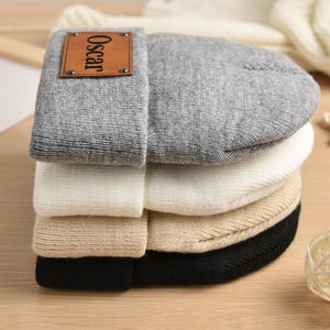 May include: A stack of four knit beanies in different colors, including gray, white, beige, and black. The top beanie has a brown leather patch with the name "Oscar" printed on it.