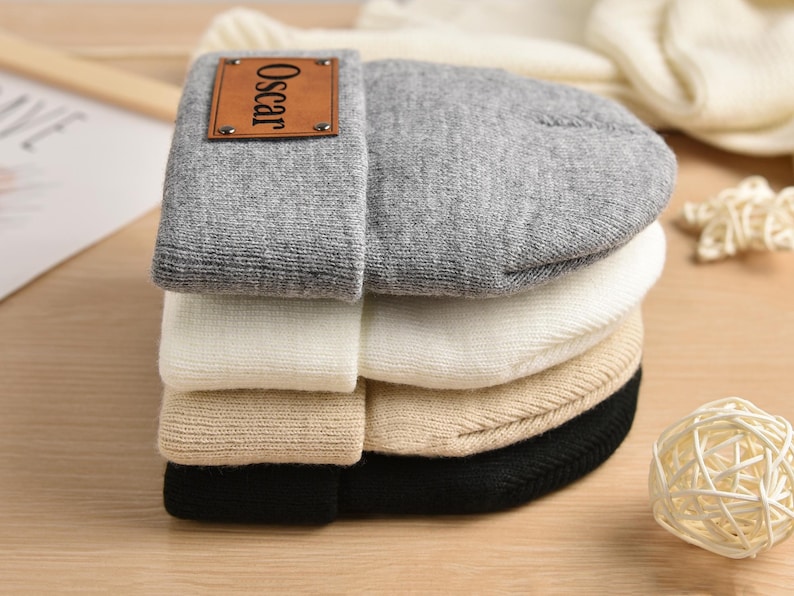 May include: A stack of four knit beanies in different colors, including gray, white, beige, and black. The top beanie has a brown leather patch with the name "Oscar" printed on it.