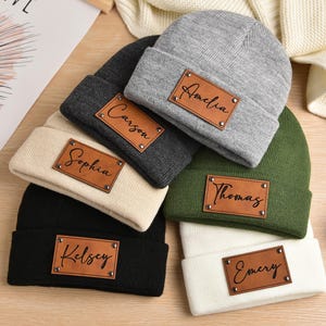 May include: Five knit beanies with personalized leather patches. The beanies are in various colors, including black, gray, white, beige, and green. The patches have the names Amelia, Carson, Sophia, Thomas, and Emery.