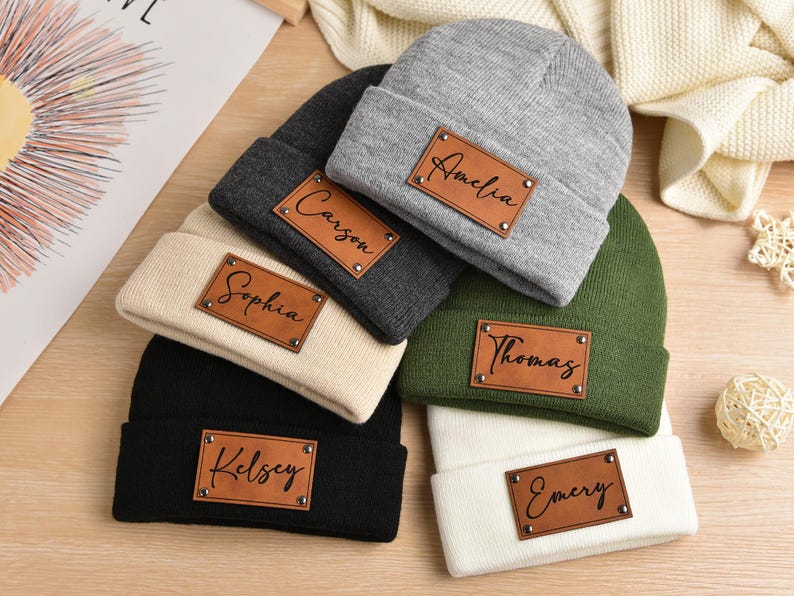 May include: Five knit beanies with personalized leather patches. The beanies are in various colors, including black, gray, white, beige, and green. The patches have the names Amelia, Carson, Sophia, Thomas, and Emery.