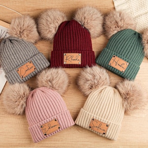 May include: A collection of ribbed knit beanies in various colors, including gray, burgundy, and green, each adorned with a faux fur pom-pom and a personalized leatherette patch. The beanies are arranged on a wooden surface.