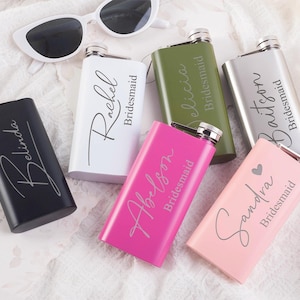 May include: A selection of personalised hip flasks in black, white, pink, green, and silver. Each flask is engraved with a name and the word "Bridesmaid". White sunglasses are in the background.