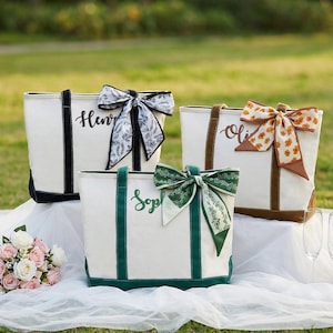 May include: Three personalized tote bags with names embroidered on them. The bags are white with black, brown, and green accents. Each bag has a decorative bow. A bouquet of flowers, champagne, and glasses are also in the image.