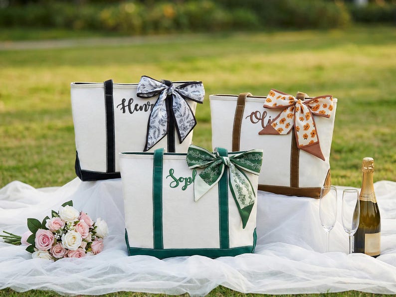 May include: Three personalized tote bags with names embroidered on them. The bags are white with black, brown, and green accents. Each bag has a decorative bow. A bouquet of flowers, champagne, and glasses are also in the image.