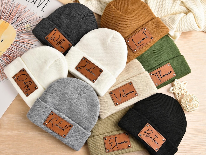 May include: Assortment of ten knitted beanies in various colors with personalized leather patches. The patches feature names such as "William", "Jackson", "Thomas", "Victoria", "Elmore", and "Bairton".