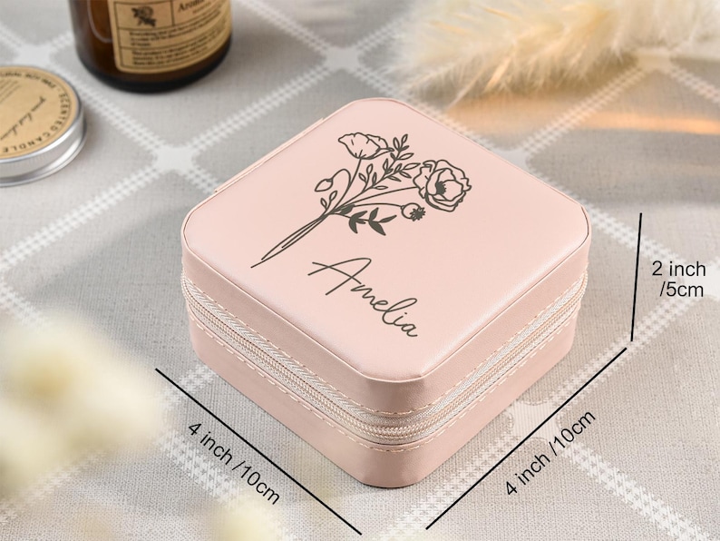 May include: A pink jewelry box with a zipper closure. The box is decorated with a floral design and the name "Amelia". The box measures 4 inches by 4 inches and 2 inches tall.