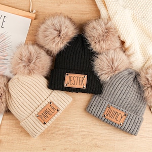 May include: Three knitted beanies with faux fur pom-poms. One is black with "JESTER" on a leather patch, another is beige with "PAMELA", and the third is gray with "QUINCY". The hats are on a wooden surface.