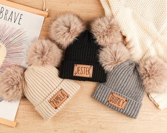 Könnte beinhalten: Drei Strickmützen mit Kunstfell-Bommeln. Eine ist schwarz mit "JESTER" auf einem Lederpatch, eine weitere ist beige mit "PAMELA" und die dritte ist grau mit "QUINCY". Die Mützen liegen auf einer Holzoberfläche.