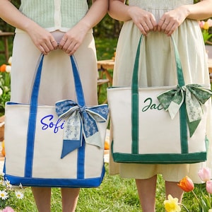 May include: Two canvas tote bags with personalized names. One bag is white with blue trim and the name "Sofi" in blue script, adorned with a blue bow. The other is white with green trim, the name "Jac" in green script, and a green bow.