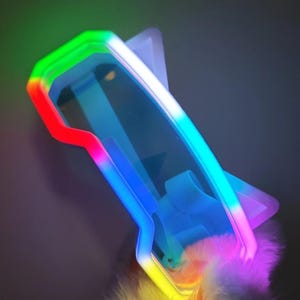 Puede incluir: Gafas futuristas con una montura de luz LED multicolor. La montura cambia de rojo a verde, a azul y a amarillo. Las gafas tienen un tinte azul claro y un acento de plumas blancas y esponjosas.