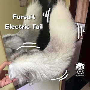 以下が含まれることがあります： ファーのスーツ用にデザインされた、黒色のベースを持つ白い毛皮の尻尾。尻尾は、ベルクロ留めの白い布製のストラップに取り付けられています。画像には「Fursult Electric Tail」というテキストが書かれています。