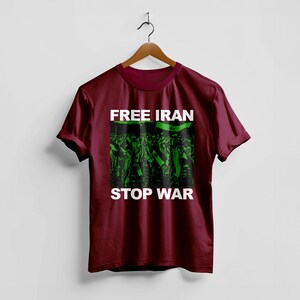 Op de afbeelding: Een bordeauxrood t-shirt met de woorden "FREE IRAN" en "STOP WAR" in witte blokletters. Het shirt heeft een groen-zwarte afbeelding van een menigte die vlaggen vasthoudt. Het shirt hangt aan een houten hanger.