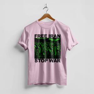 Op de afbeelding: Een lichtroze t-shirt met de woorden "FREE IRAN" en "STOP WAR" in zwarte letters. Het shirt heeft een groen-zwarte afbeelding van een menigte met vlaggen. Het t-shirt hangt aan een houten hanger.