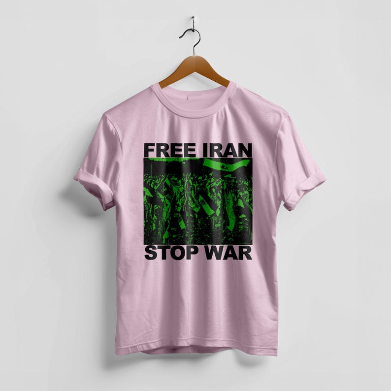 Op de afbeelding: Een lichtroze t-shirt met de woorden "FREE IRAN" en "STOP WAR" in zwarte letters. Het shirt heeft een groen-zwarte afbeelding van een menigte met vlaggen. Het t-shirt hangt aan een houten hanger.