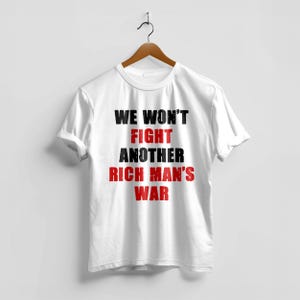 May include: White t-shirt with the text "WE WON'T FIGHT ANOTHER RICH MAN'S WAR" in black and red. The shirt is hanging on a wooden hanger against a white background. The text is centered on the front of the shirt.