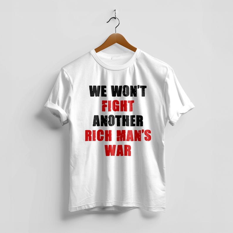 May include: White t-shirt with the text "WE WON'T FIGHT ANOTHER RICH MAN'S WAR" in black and red. The shirt is hanging on a wooden hanger against a white background. The text is centered on the front of the shirt.