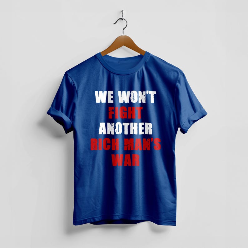 May include: A blue t-shirt with the text "WE WON'T FIGHT ANOTHER RICH MAN'S WAR" in white and red lettering. The shirt is hanging on a wooden hanger against a white background.