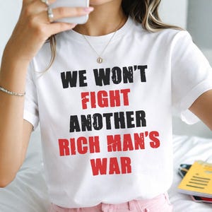 May include: White t-shirt with the text "WE WON'T FIGHT ANOTHER RICH MAN'S WAR" in black and red lettering. The shirt is short-sleeved and worn by a person. The person is wearing pink shorts and holding a phone.
