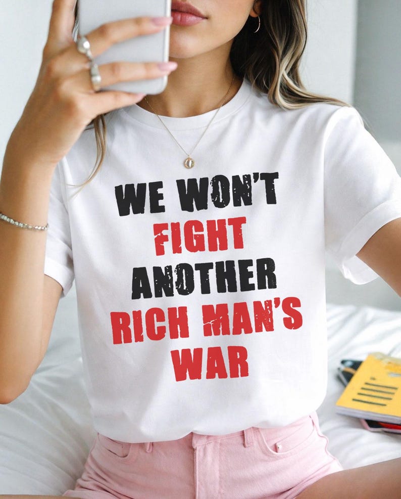 May include: White t-shirt with the text "WE WON'T FIGHT ANOTHER RICH MAN'S WAR" in black and red lettering. The shirt is short-sleeved and worn by a person. The person is wearing pink shorts and holding a phone.
